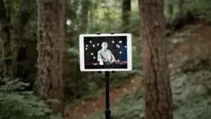 An ipad on a stand is in a forest setting between two trees. On the screen is a man being interviewed