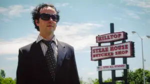 A man in a suit and large sunglasses stands facing the camera. Behind him is a sign for the Hilltop Steakhouse